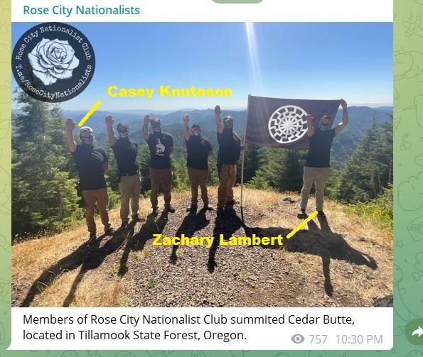 Neo-Nazi group Rose City Nationalists posing in a clearing on a mountain. They are all wearing skull masks and sieg heiling, and Zachary Lambert is helping to hold a sonnenrad flag.
