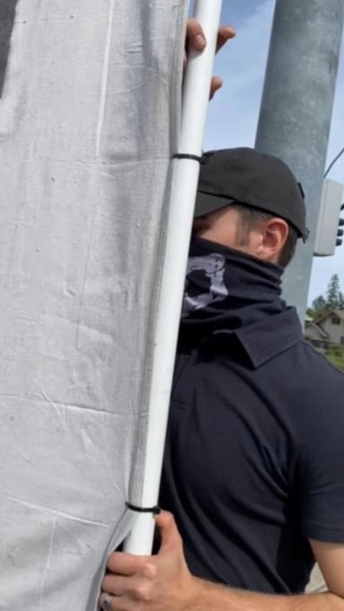 Neo-Nazi Zachary Lambert wearing a skull mask and holding a banner that says "gender theory is poison". He is attempting to hide his face behind the banner as someone films him.