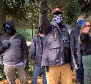 A group of neo-nazis at a protest. Three of them look bored, and one is sieg heiling for the camera. Dixie Bailey is standing with them giving a middle finger.