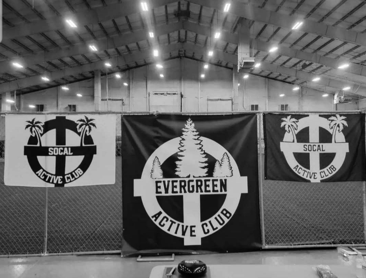 B&W Image from the Martyrs Day Rumble in Pasco. Shows the inside of the Hapo Center with a chain link fence on which three flags are hung: two small SoCalAC flags and one large EAC flag in the center
