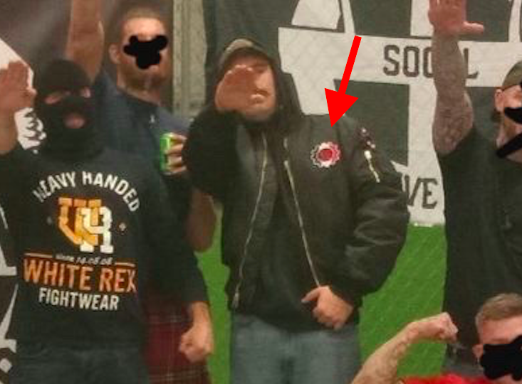 Patrick Mushaney and other nazis all sieg heiling at the martyrs day rubmle in pasco. There is a Hammerskins patch on Mushaney's bomber jacket, with a red arrow added to highlight it.