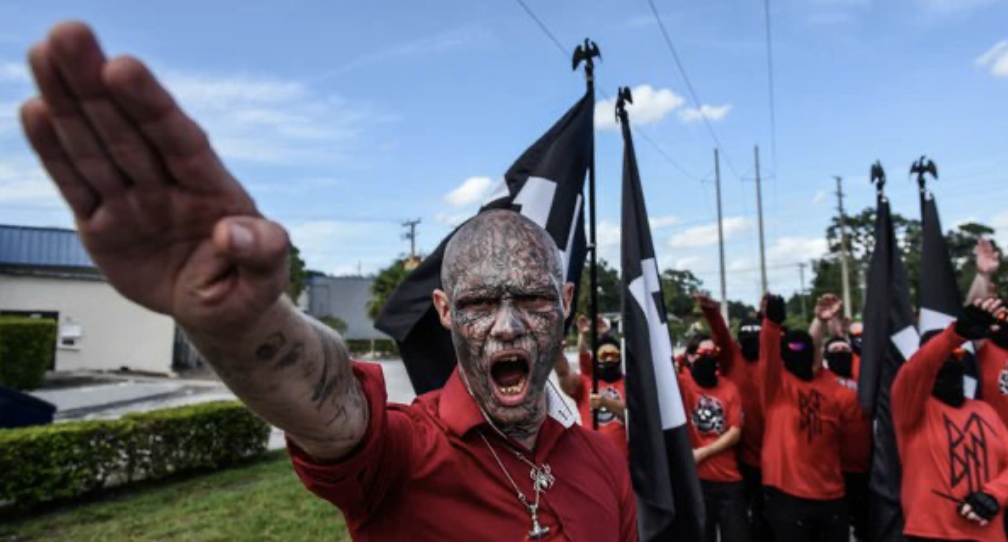 Kent McClellan, aka Boneface, raises his right arm in a nazi salute with mouth wide open. Kent's face and head are completely covered in tattoos. There is a group of nazis in red shirts and black face masks throwing nazi salutes behind him.