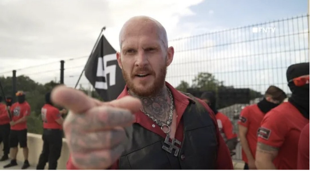 Chris Pohlhaus, aka Hammer, points at the camera. He is wearing a swastika necklace, red button down shirt that is open at the top and exposing a ram's head tattoo. There is a swastika flag flying behind him where a group of nazis are standing wearing black face masks and red shirts.