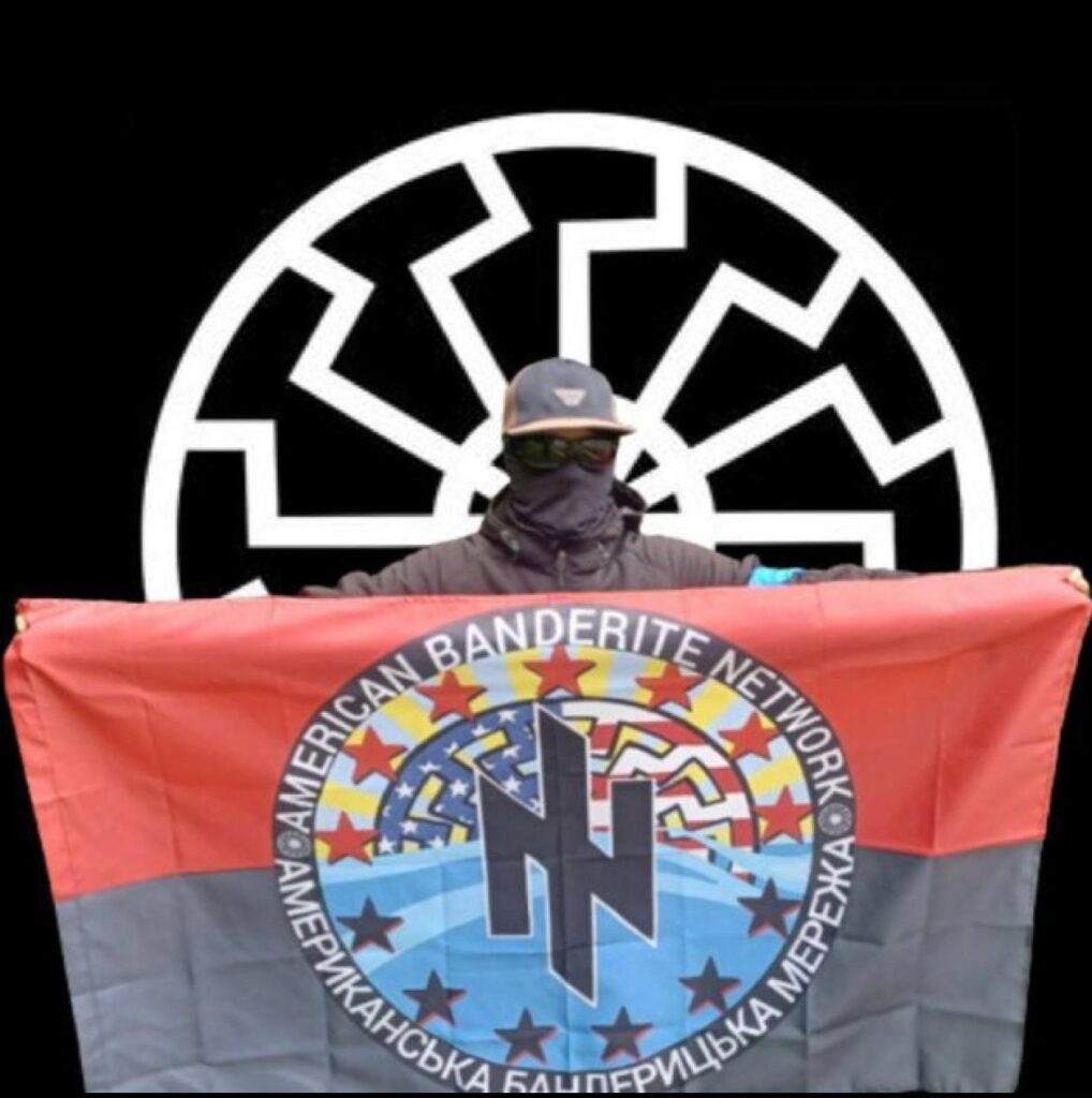 A young man wearing a black face gaiter, sunglasses, baseball hat, and black coat with blue painters tape on the arm holds up a flag representing the American Banderite Network. There is a sonnenrad behind him.