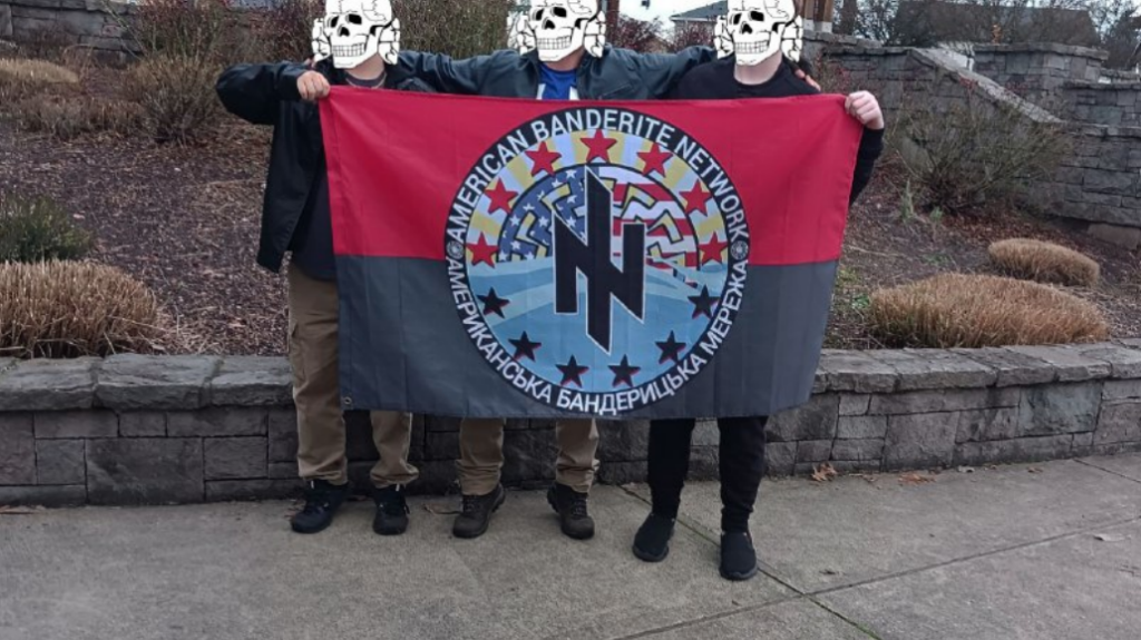 3 teenagers stand closely to one another while holding an American Banderite Network flag. Their faces are covered to conceal their identities