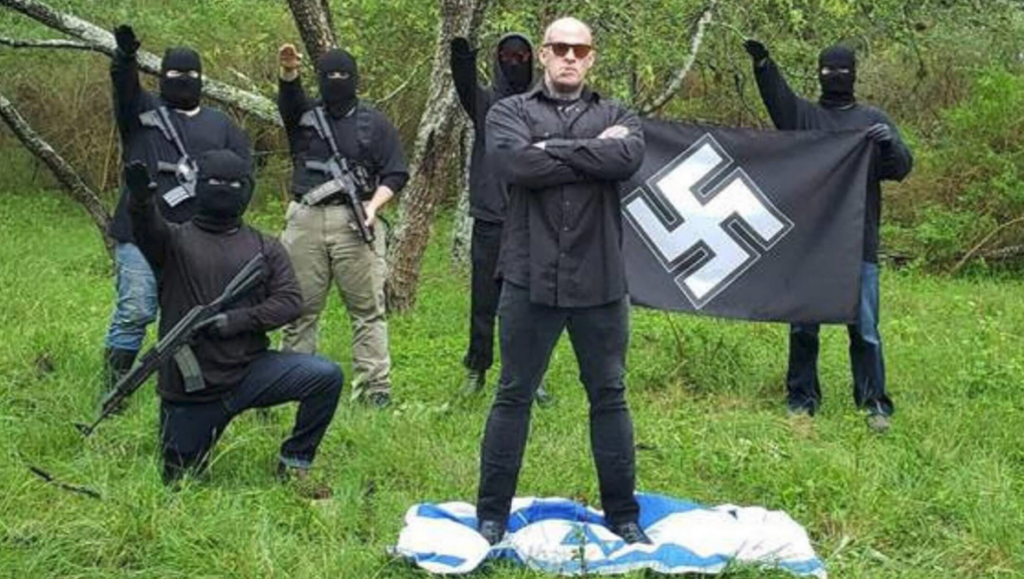 Chris Polhaus posing in front of a group of masked nazis holding rifles and doing nazi salutes, and holding a swastika banner.