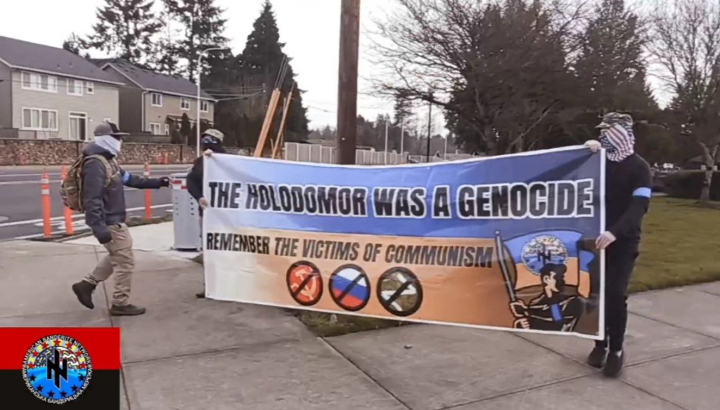 Three masked boys holding a banner at a park that says "Holodomor was a genocide, remember the victims of communism" and has nazi imagery on it
