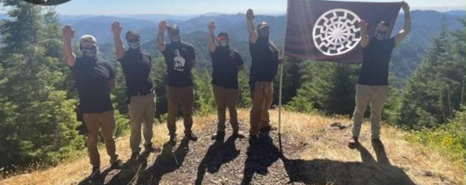 6 members of Rose City Nationalists on a ridge in the woods, holding a sonnenrad flag and doing nazi salutes