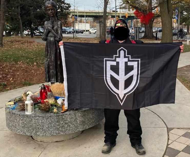 Pic of a masked Andrew Benson at a memorial holding a fascist banner