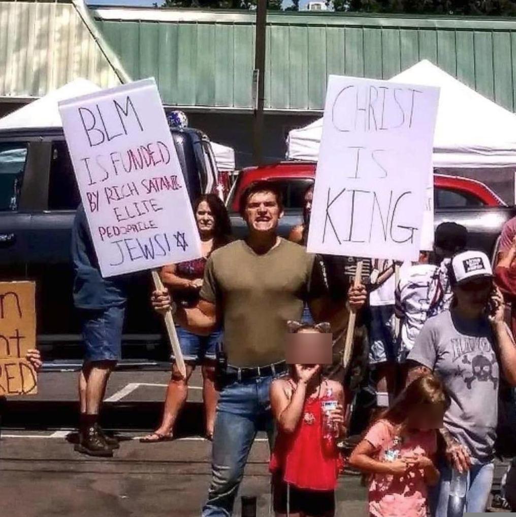 A young adult white male carrying a gun stands in a parking lot with a group of people who include young children. He holds a sign in one hand that reads, "BLM is funded by rich satanic elite pedophile Jews!" In the other hand he holds a sign that reads "Christ is King".