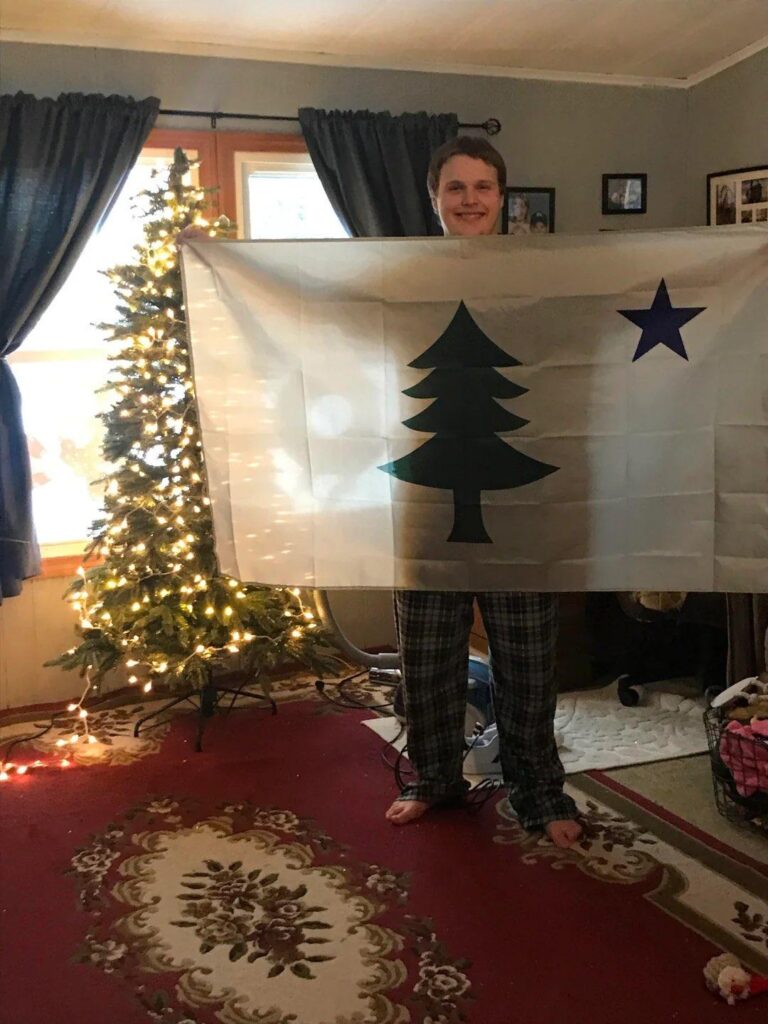 A smiling Ian, barefoot and in pajamas, holds up an old version of the Maine flag. It has a pine tree in the center and a north star in one corner. He is standing next to a Christimas tree.