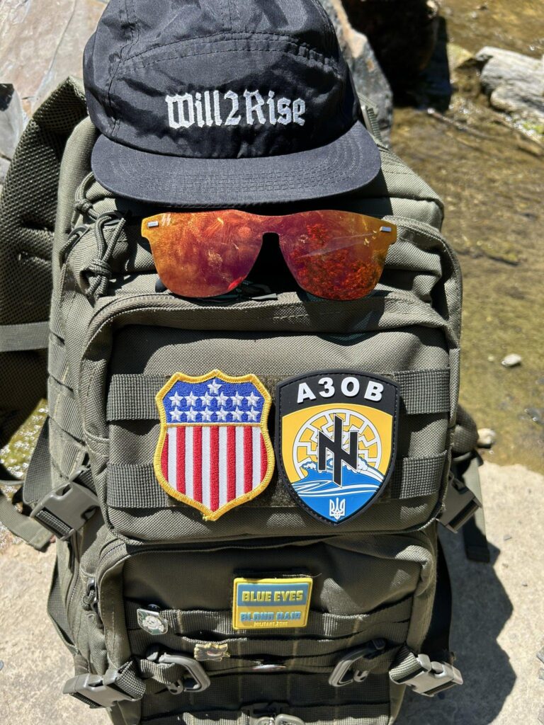 A backpack is placed on the ground next to a stream. It has multiple patches on it, including one for Azov. There is a white nationalist "Will 2 Rise" ballcap placed on the top of the pack. And a pair of Hammershades are placed just under the brim.