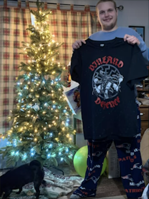 Ian stands next to a Christmas tree holding up a tshirt with nazi symbols and smiling. He has presumably received the shirt as a Christmas gift.